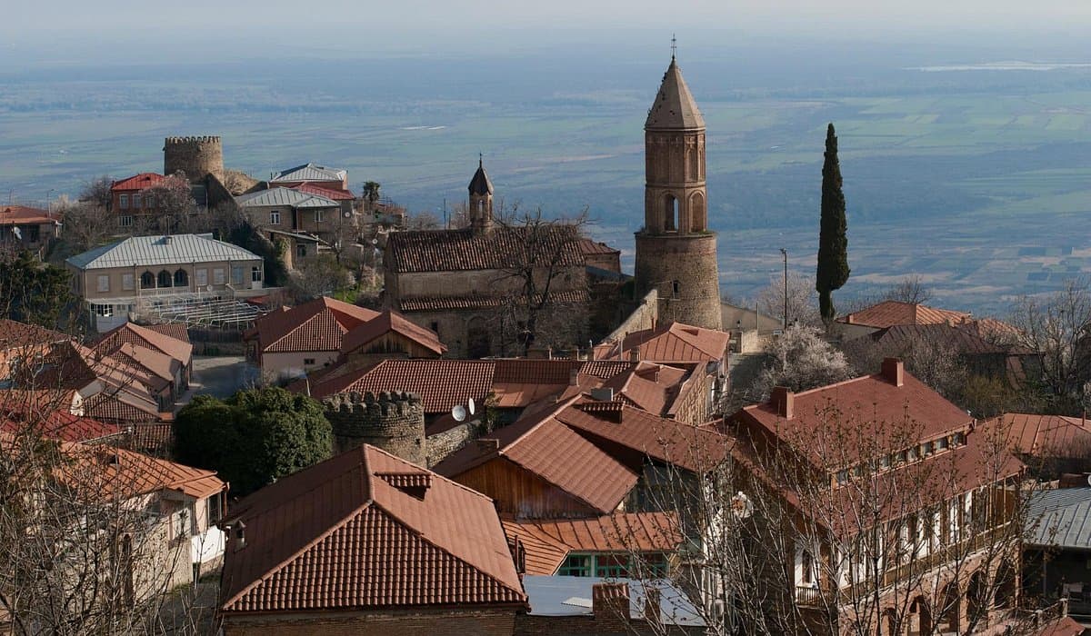 Rooftops of Sighnaghi spread across a hilltop in Kakheti, with a round stone bell tower and fortress walls rising above the Alazani valley plain.
