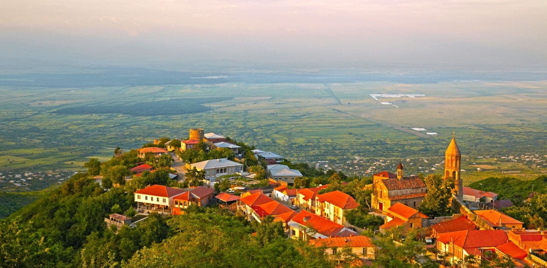 Hilltop town of Sighnaghi with red-roofed stone buildings, a church tower, and the vast Alazani valley stretching below at golden hour.