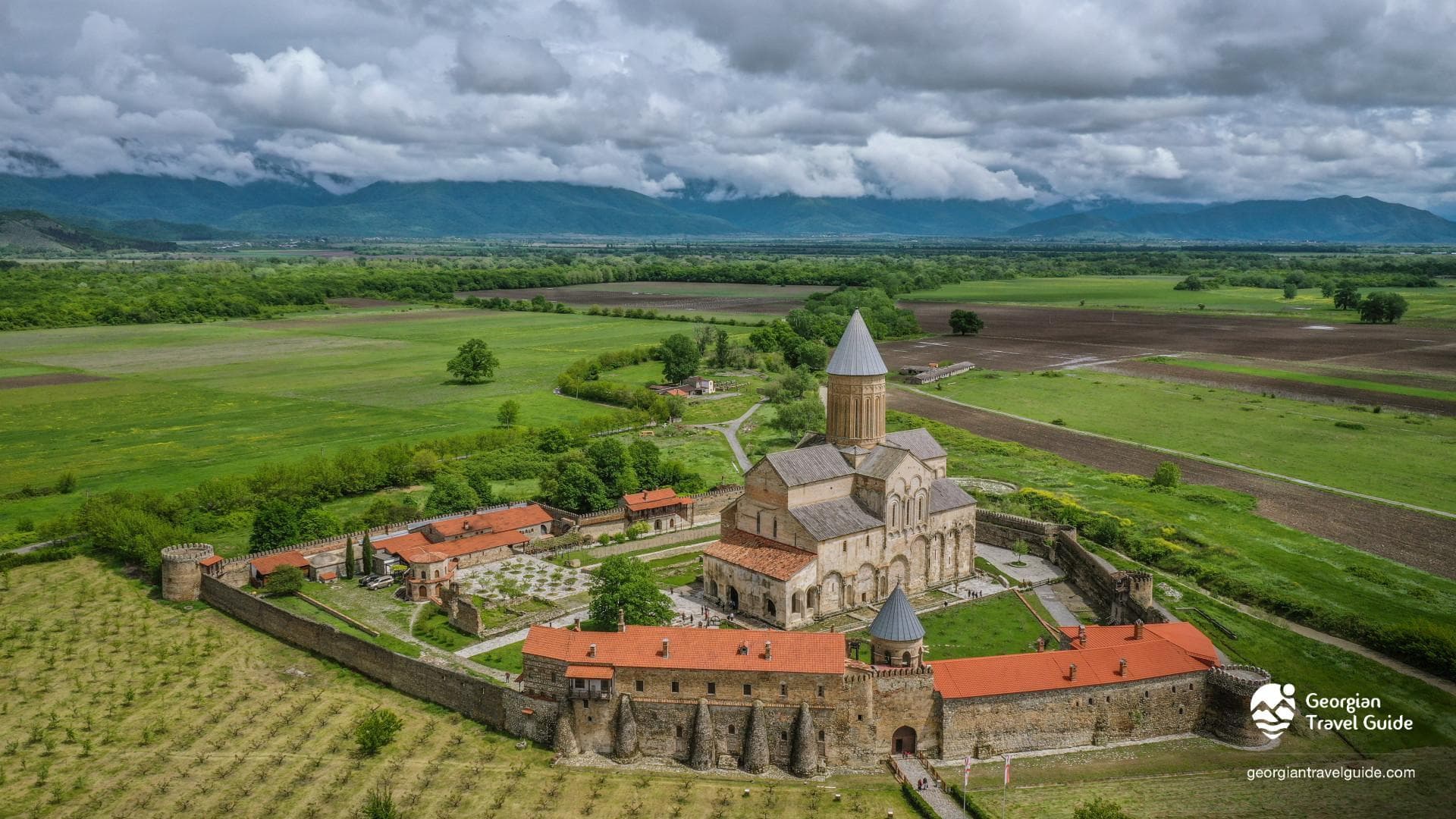 Aerial view of Alaverdi Cathedral in Kakheti, surrounded by stone walls, red-roofed buildings, green fields, and the Caucasus mountains under a cloudy sky.